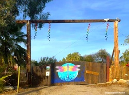 Front gate of DragonflyHill. A large wooden gate with a square arch made of reclaimed utility poles. The gate has a mural of a dragonfly on it and there are rainbow streamers hanging from the arch.